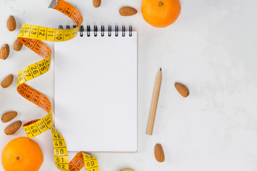 Diet concept - notebook, vintage orange yellow measuring tape, mandarins, almonds, pencil on white concrete background. Top view, flat lay, copy space, mock-up