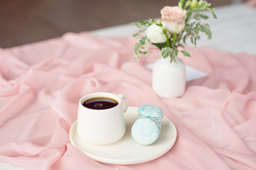 French macaroon blue plate on the pink and coffee cup standing on a wooden table with a pink tablecloth white vase with flowers roses and greens.