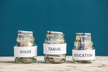 Jars with money for different needs on table against color background