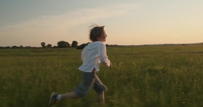 Happy child running on the meadow in the summer while sunset. Happy 7 year old cheerful boy runs and looks back, on field. Happiness concept. Active smiling boy on nature in spring. Slow motion