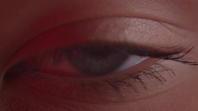 Close-up shoot of light-blue eye blinking with reflection of lamp on it in red light.