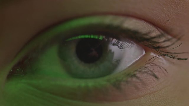 Close-up shoot of light-blue eye blinking with reflection of lamp on it in green light.