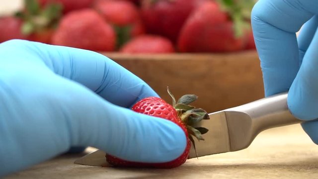 close-up of a girl's hands in blue rubber gloves cutting a strawberry with a silver metal knife on a wooden board against a blurred image of a wooden plate with strawberries