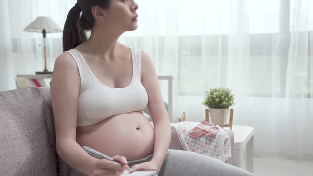 low to high angle of young asian pregnant woman with bare belly sitting on couch with baby clothes beside. beautiful future parent relax on sofa writing on notebook and looking window daydreaming