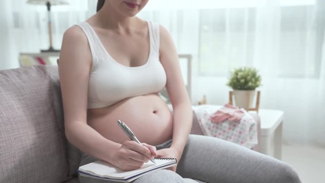 unrecognized young healthy pregnant woman in sports bra and leggings sitting on sofa in home living room. asian lady motherhood writing in notebook with pen while relax on couch in cozy bright house.