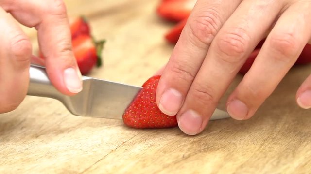 close-up of a girl's hands cutting a strawberry with a silver metal knife on a wooden board against a blurred image of a wooden plate with strawberries