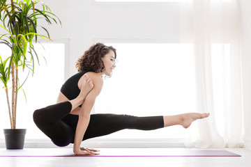 Young brunette female is balancing at leg over shoulder pose. Athletic woman in black uniform is practicing yoga, meditating in eka hasta bhujasana posture at fitness studio. Healthy lifestyle concept