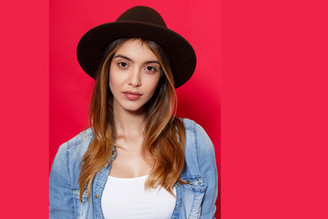 Close up of a hot brunette girl in hat and denim shirt, looking with attitude on camera, isolated on red background.