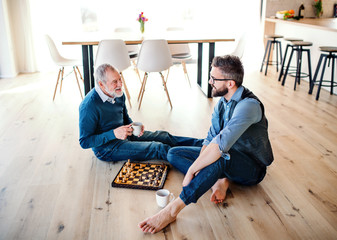 An adult hipster son and senior father sitting on floor indoors at home, playing chess.