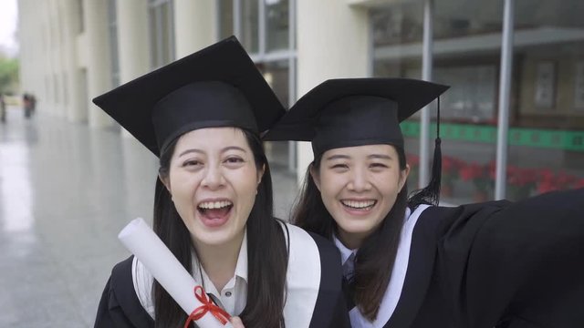 slow motion happy group of mature students on graduation day pose for selfie. two asian best girl friends in academic dress and caps taking self picture. smiling attractive female hugging face camera