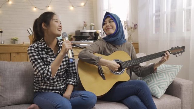 Happy young international multi friends in shared apartment playing guitar singing with microphone. music asian girls roommates relaxing in dormitory kitchen sitting on couch having fun smiling.