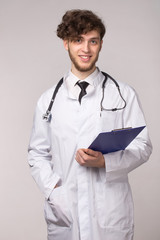 Portrait of smiling confident young smiling medical doctor with sthetoscope and clipboard over light gray background