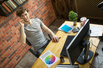 Young smiling digital artist designer talking by smartphone with his customer at his workplace in loft style office.
