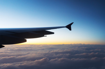 Wing of airplane flying above the clouds in sky with sunset