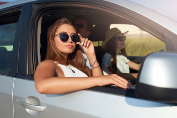 Group of friends rented a car on summer road trip and arrived to the sea beach. Woman in glasses looks out of the car window. Passanger girl having fun with friends in vehicle. Travel lifestyle.