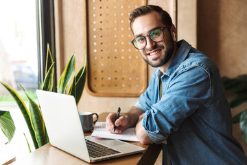 Photo of attractive smiling man writing and using earpod with laptop while working in cafe indoors