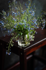 Forget-me-not's bouquet in small glass jar on wooden stool, dark background