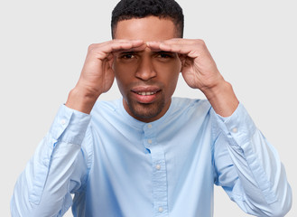 Closeup studio portrait of young African American man posing for advertisement wearing blue shirt, looking away with hands on forehead, isolated on white wall. People, emotion