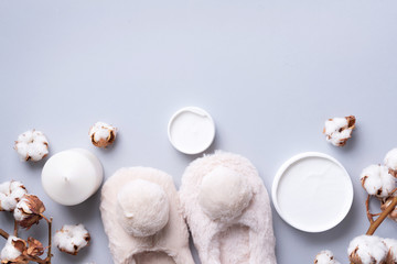 Bathroom accessories, nude fluffy home slippers, cotton flowers, candle, skin care products on grey background with copy space. Top view. Copy space. Spa and body treatment concept.