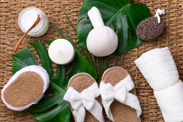 Spa, wwellness, skin care, beauty and relax concept. Spa tools: white towel, bamboo slippers, herbal ball, cream, wooden brush, coconut oil, monstera on rattan background.