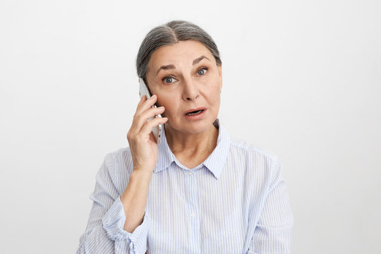 Isolated image of emotional astonished elderly female wearing blue shirt keeping mouth wide opened while receiving shocking news talking on mobile phone, lost for words, can't say anything