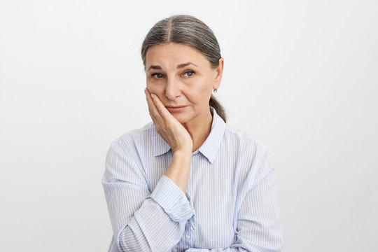 Frustrated female pensioner in blue shirt holding hand on her face having worried uneasy facial expression, thinking about her future. Unhappy sad senior woman suffering from terrible toothache