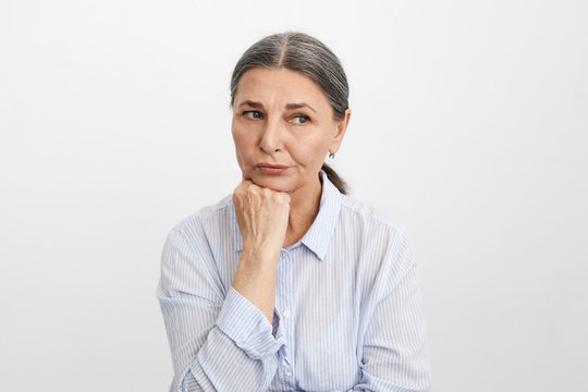 Studio picture of upset frustrated elderly senior European female wearing blue striped shirt placing hand under chin, thinking about her children who don't visit her, feeling lonely and unhappy