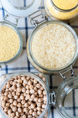 Various raw grains, flakes and legumes in glass jars, top view. Uncooked chickpeas, polenta, couscous and rice on a white checkered tablecloth.