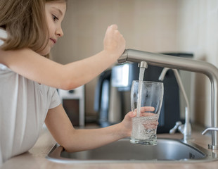 Portrait of a little caucasian girl gaining a glass of tap clean water. Kitchen faucet. Cute curly kid pouring fresh water from filter tap. Indoors. Healthy life concept