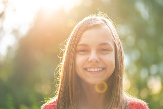 Close up portrait of teenage girl with sun rays filtering through her hair. Happy smiling teen at summer park in flare sunshine. Child looking at camera during sunset.