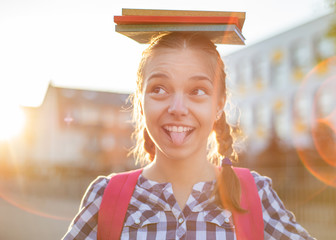 Close up portrait of cheerful teenage girl with backpack and books on head in sun rays. Happy smiling teen with bag go to school with fun or returning home in flare sunshine. Child showing tongue