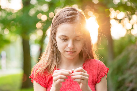 Portrait Of Teenage Girl With Sun Rays Filtering Through Her Hair. Happy Smiling Teen Guesses On A Chamomile  At Summer Park In Flare Sunshine. Child Tears Off The Petals Of Daisy During Sunset.