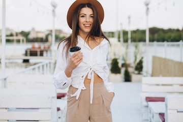 Fototapeta premium A stylish woman drinking coffee and walking along the beachfront in a warm summer day on the sunset