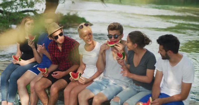  A group of young friends enjoying watermelon while sitting on the wooden bridge over the river in beautiful nature