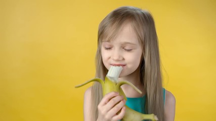 Portrait of cute little girl standing isolated on yellow background and smiling while eating banana and looking at camera