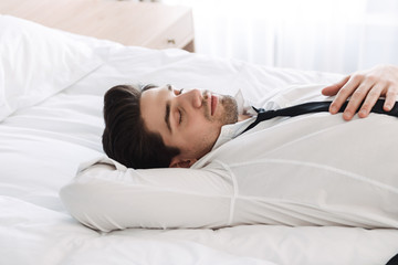 Photo in profile of calming bearded businessman lying with eyes closed on bed in hotel apartment