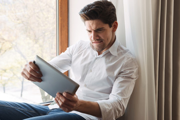 Photo of excited focused man playing game on tablet computer while sitting at window in living room