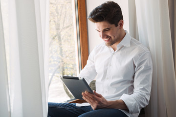 Photo of smiling brunette man making video call on tablet computer while sitting at window in living room