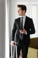 Photo of satisfied young businessman holding cellphone and luggage in hotel apartment