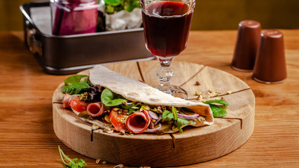 Concept Italian cuisine. Piadina with ham, tomatoes, mix lettuce, pistachios, cucumbers on wooden board. Glass of red wine on the table. Beautiful serving dishes in the restaurant.