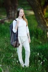 young woman smiling in white clothes came out on a glade with dandelions to the lake to do yoga with a rug in a bag behind her back