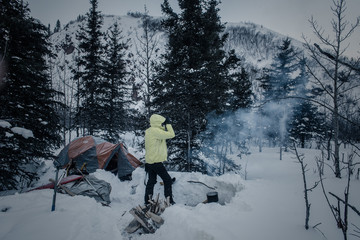 Woman in light green jacket by morning campfire and tent in deep snow in winter mountains