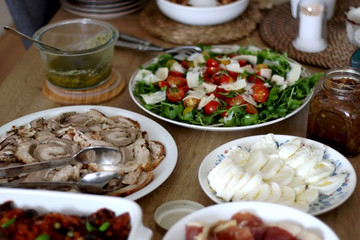 Lavish homemade lunch served on table: focaccia bread, prosciutto, porchetta, mozzarella cheese, pesto, caponata, caramelized onion, with arugula and cherry tomato salad. Selective focus.