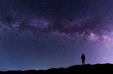A silhouette stands on in the hill and looks at the Milky Way galaxy.