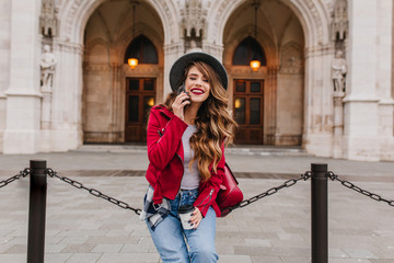 Fashionable girl with long hairstyle talking on phone near amazing historical building in weekend. Outdoor photo of brunette caucasian lady with cup of tea posing on beautiful architecture background.