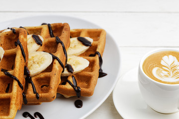 Closeup homemade belgian waffles with banana slices topped with chocolate and cup of coffee on white wooden table. Angle view with shallow focus.