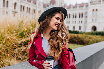 Fototapeta premium Joyful long-haired woman with trendy black manicure looking away while waiting someone near old building. Photo of pretty caucasian female model in hat drinking coffee outdoor.