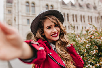 Joyful caucasian girl spending vacation in Hungary and making selfie. Outdoor portrait of funny elegant lady in black hat taking picture of herself, walking past old building.