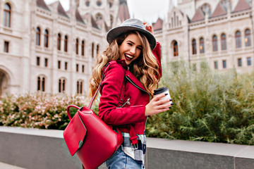 Shapely girl with wavy hair laughing while exploring old part of city. Charming female tourist in...