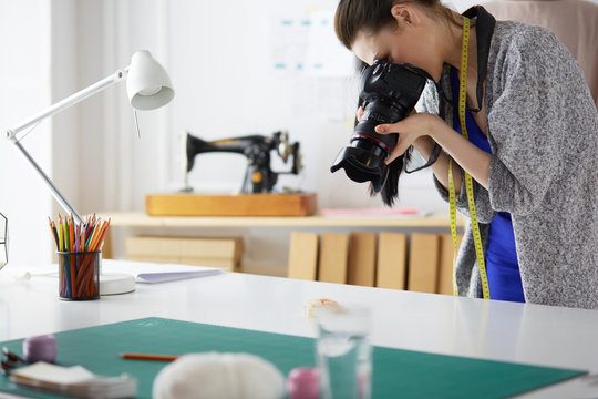 Young designer woman taking photos with digital camera while standing near the desk in her workshop